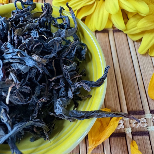 Dark, twisted Jiang Hua (Ginger Flower Fragrance) Phoenix Dan Cong oolong tea leaves in a yellow bowl next to yellow flowers and petals
