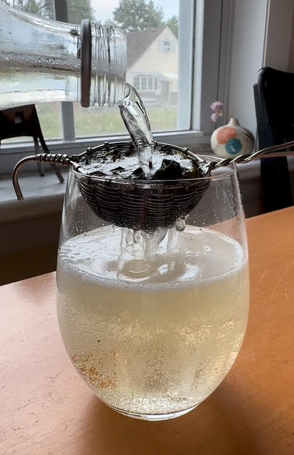 Glass bottle pouring sparkling tea through a silver strainer into a stemless wine glass with ice cubes. The tea is fizzing.