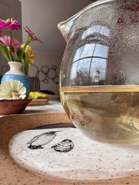 Glass, rounded fairness pitcher with pale yellow Chinese yellow tea sitting on a clay spoon rest with painted penguins