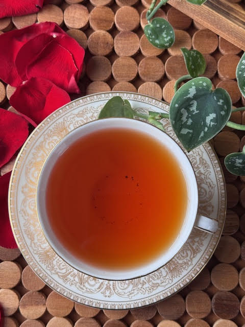 Rose Label Gold 2025 white and gold teacup with a saucer. Red rose petals and green ivy are nearby