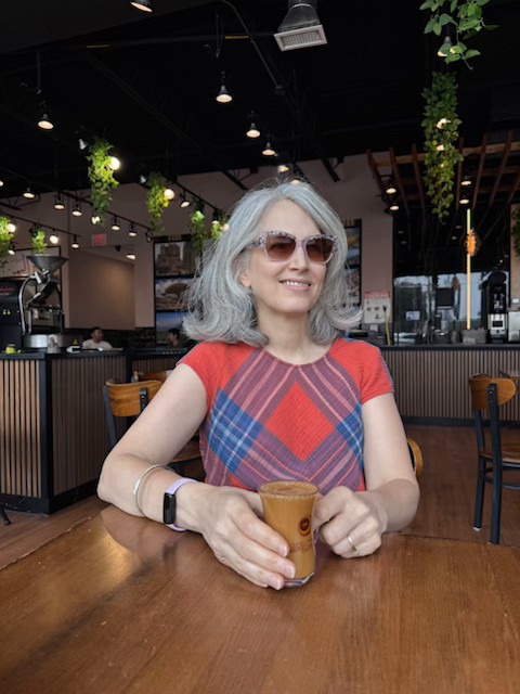 Photo of Traci Levy, aka Tea Infusiast--a white woman with silver hair, wearing sunglasses and a red shirt with blue plaid. Her hands rest on a curved, glass teacup full of Adeni Chai at Qahwah House