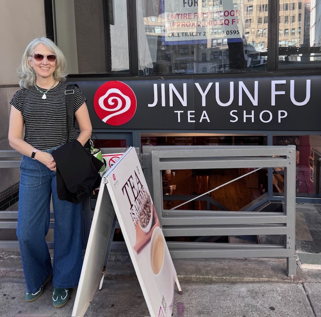 Traci (a.k.a. Tea Infusiast) wearing a black shirt with white stripes and jeans and sunglasses in front of the sign for "Jin Yun Fu Tea Shop"