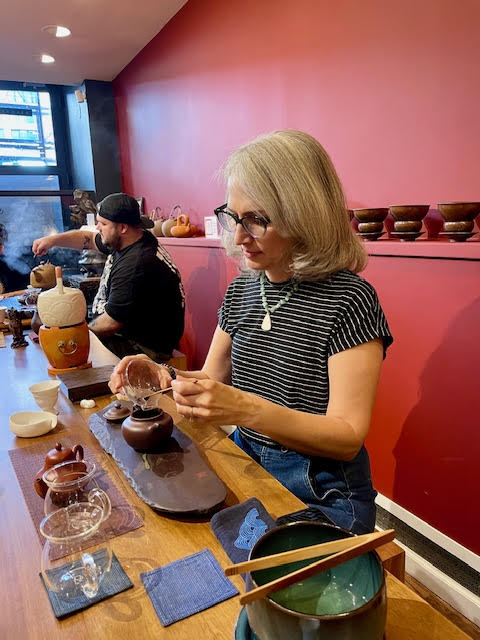 Traci Levy--a white woman with silver hair and green glasses wearing a black t-shirt and sliding tea into a clay teapot at Jin Yun Fu