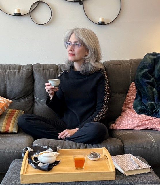 Photo of Traci --a white woman with shoulder length silver hair wearing glasses and all black and holding a teacup. This image symbolizes her monthly event for TIN subscribers--Tea Mindful Together
