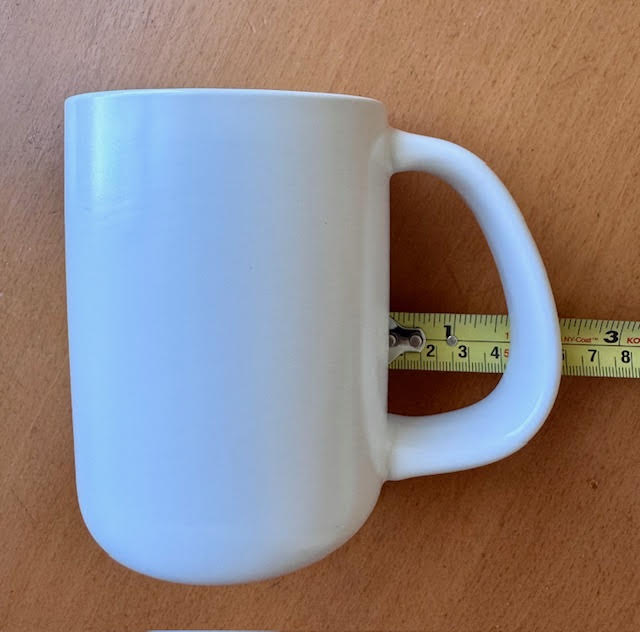 side view of white accessible mug by CURVD with measuring tape showing the distance between the handle and the wall of the mug