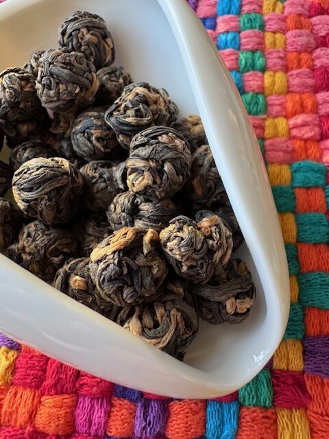 tea leaves rolled into spheres--with dark and caramel brown tea leaves. The leaves are in a white dish on a very colorful piece of fabric