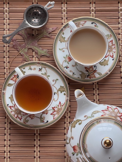 two white teacups with Oberon Wedgwood pattern--one cup has plain black tea, the other has tea with milk. The teacups are next to a silver strainer and a teapot