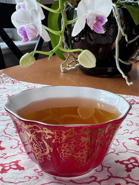 Dark amber colored steeped burdock root tea in a red teacup with gold scrolling and a white interior. A white and fuschia orchid is blooming in the background