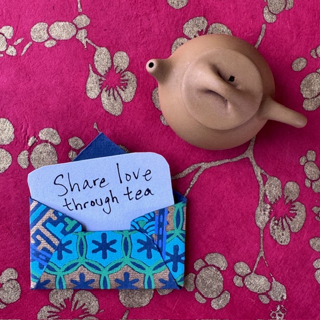 Blue and gold patterned envelope with a white piece of paper coming out that says "share love through tea." A small clay teapot sits next to the envelope.