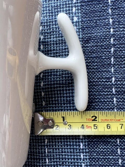 Close up of an accessible T-shaped handle (T Handle) mug's handle next to a yellow measuring tape showing the distance between the side of the mug and the bottom of the handle.