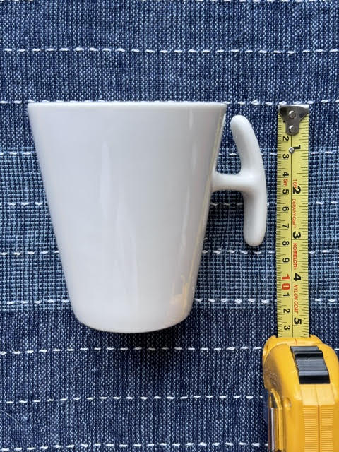 White porcelain mug with a T handle next to a yellow measuring tape measuring the height of the mug.