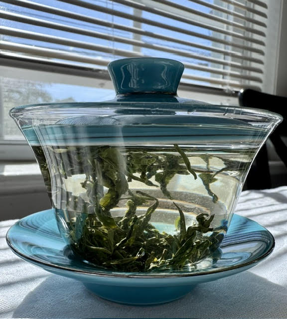 Glass gaiwan with a turquoise blue lid and base with water and green tea leaves. Glass teaware is helpful to enjoy drinking less tea.