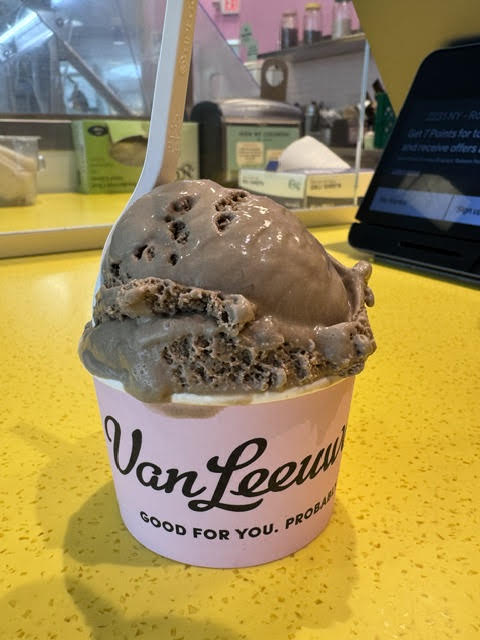 Scoop of dark brown Hojicha tea ice cream at Van Leeuwen in a pink cup with a spoon sticking out of the scoop.