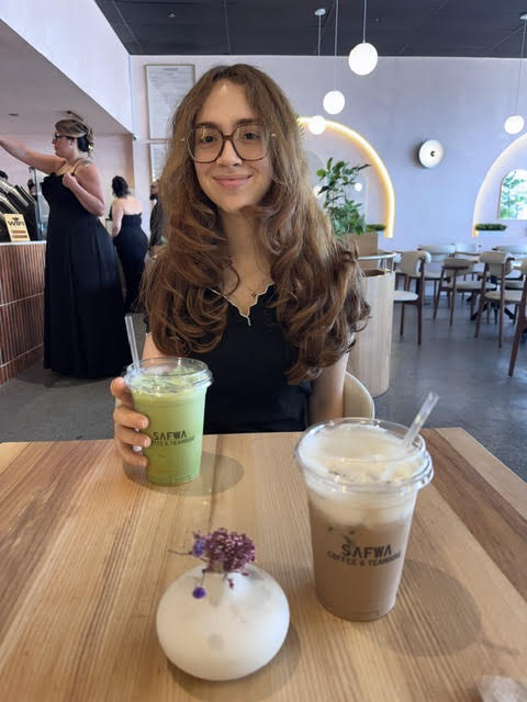 Woman with long ginger brown hair and glasses holding a pistachio matcha at smiling at Safwa Coffee & Teahouse in Westbury, NY
